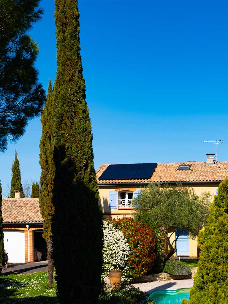 Panneaux solaires sur le toit d'une maison, avec un grand jardin ayant plusieurs arbres, ainsi qu'une piscine. Ciel dégagé et environnement ensoleillé