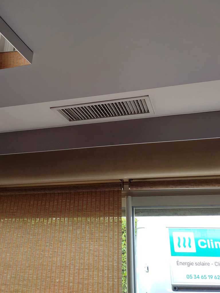 Grille de ventilation d’un système de climatisation gainable installée au plafond, dans une pièce lumineuse avec stores en rotin