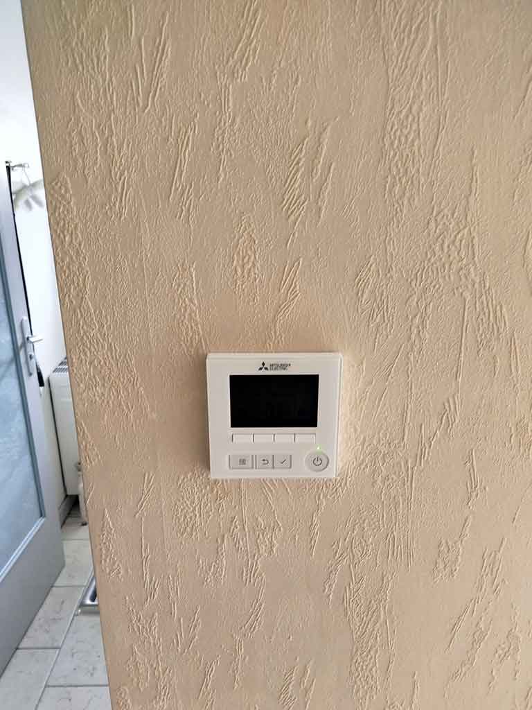 Thermostat mural Mitsubishi Electric pour climatisation gainable, fixé sur un mur beige texturé, pour réguler la température intérieure